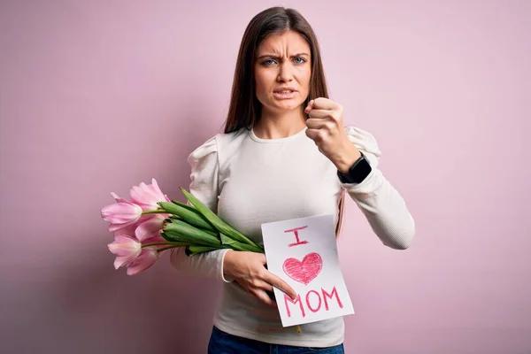 Young beautiful woman with blue eyes holding love mom message and tulips on mothers day annoyed and frustrated shouting with anger, crazy and yelling with raised hand, anger concept