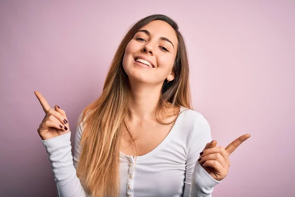 ピンクの背景に白いTシャツを着た青い目をした若い美しいブロンドの女性は さまざまな方向への指で自信を持って指して笑っています 広告のコピースペース — ストック写真