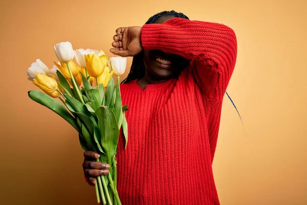 Young african american plus size woman with braids holding bouquet of yellow tulips flower Smiling cheerful playing peek a boo with hands showing face. Surprised and exited