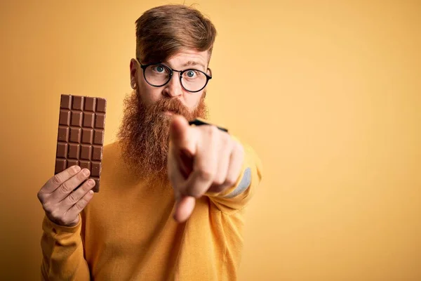Irish redhead man with beard eating a sweet chocolate bar over yellow isolated background pointing with finger to the camera and to you, hand sign, positive and confident gesture from the front