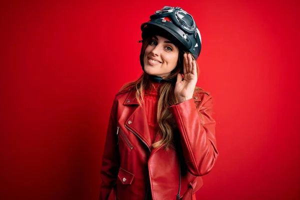 噂やゴシップに耳を傾ける耳の上に手で笑みを浮かべて 赤の背景に元ヘルメットを身に着けている若い美しいブルネットのオートバイの女性 病気の概念 — ストック写真