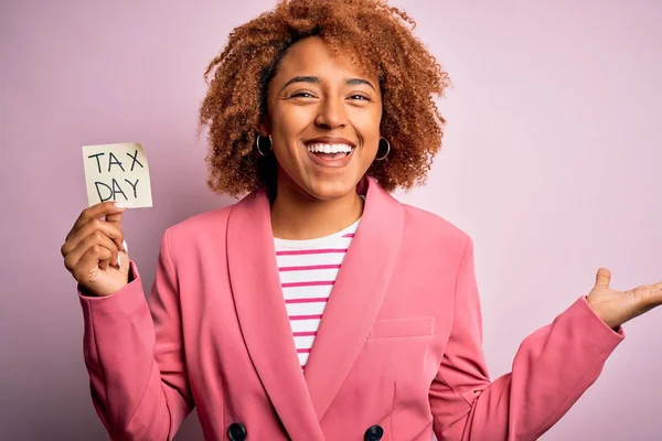 Young African American afro woman with curly hair holding papaer with tax day message very happy and excited, winner expression celebrating victory screaming with big smile and raised hands