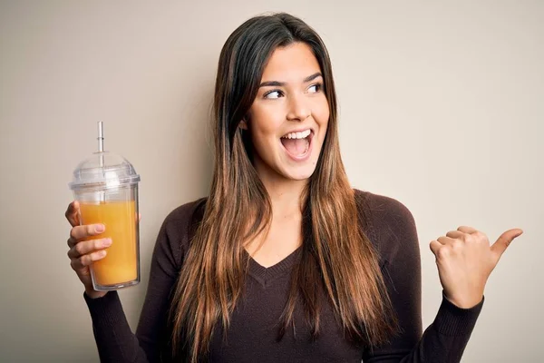 年轻美丽的姑娘喝着一杯健康的橙汁 隔着孤立的白色背景 指指点点 笑容满面地站在一旁 — 图库照片