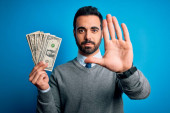 Mladý pohledný muž s plnovousem drží hromadu dolarů bankovky nad modrým pozadím s otevřenou rukou dělat stop znamení s vážným a sebejistý výraz, obrana gesto