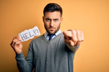 Çok çalışan genç iş adamı, sarı zemin üzerinde kağıt tutarken kamerayı işaret ederek yardım istiyor ve sen, el işareti, ön taraftan pozitif ve kendinden emin bir hareket.