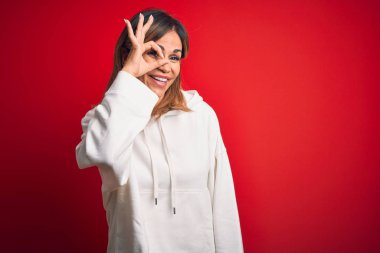 Orta yaşlı güzel sporcu kadın izole edilmiş kırmızı arka plan üzerine gündelik eşofman giyip el hareketleriyle gülümsüyor ve mutlu yüzle parmaklarının arasından bakıyor..