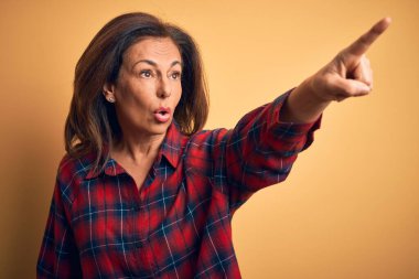 Orta yaşlı, güzel bir kadın, sıradan bir gömlek giymiş, sarı arka planda dikiliyor, parmağı önde, ağzı açık, şaşırmış, ön yüzünde bir ifade var.