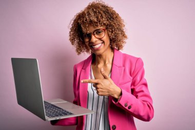 Kıvırcık saçlı, pembe arka planda dizüstü bilgisayar kullanan Afro-Amerikan iş kadını elini ve parmağını göstererek mutlu bir şekilde çalışıyor.