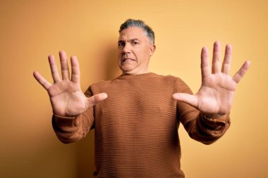 Orta yaşlı, yakışıklı, gri saçlı, sarı arka planda rahat süveter giyen, korku ifadesinden korkan ve dehşete düşmüş bir adam. El hareketlerini bırak, şok içinde bağır. Panik konsepti.