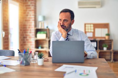 Orta yaşlı yakışıklı iş adamı ofiste dizüstü bilgisayar kullanarak oturmuş kollarını kavuşturup depresyondan sıkılmış ve yorgun göründüğünü düşünüyor..