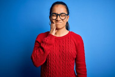 Genç ve güzel Asyalı bir kadın diş ağrısı ya da diş ağrısı yüzünden mavi arka planda gözlük takıyor ve dudaklarına acı dolu bir ifadeyle dokunuyor. Dişçi