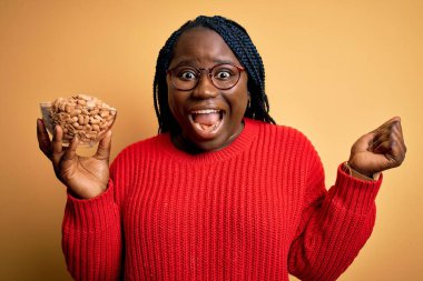 Young african american plus size woman with braids holding bowl with healthy peanuts screaming proud and celebrating victory and success very excited, cheering emotion clipart