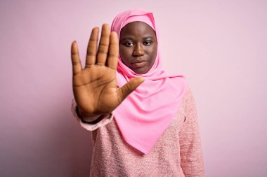 Genç Afro-Amerikan artı cüsseli kadın soyutlanmış pembe arka plan üzerine Müslüman başörtüsü takıyor ve avuç içiyle şarkı söylemeyi kesiyor. Yüzünde olumsuz ve ciddi bir jest olan uyarı ifadesi.