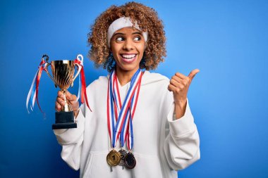 Genç Afro-Amerikan spor yapan, spor yapan, madalya kazanan, kupa kazanan ve gülümseyen bir gülümsemeyle baş parmağıyla yüzünü gösteren bir kadın.