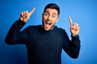 İzole edilmiş mavi arka plan üzerinde duran sıradan bir süveter giyen genç yakışıklı. Şaşırmış, şaşırmış ve parmaklarıyla işaret eden ve kollarını kaldıran..