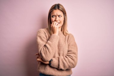 Pembe yünlü arka plan üzerine kışlık yün kazak giyen genç, güzel sarışın kadın elleri ağız yiyen tırnaklarla gergin ve stresli görünüyor. Endişe sorunu.