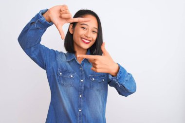 Young beautiful chinese woman wearing denim shirt standing over isolated white background smiling making frame with hands and fingers with happy face. Creativity and photography concept.