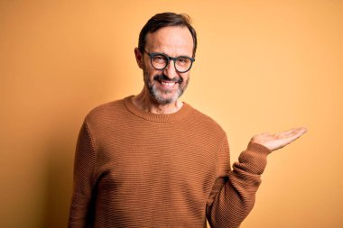 Kahverengi süveter ve gözlük takan orta yaşlı beyaz bir adam izole edilmiş sarı arka planda mutlu bir şekilde gülümseyen ve avuç içi kameraya bakan bir adam..