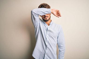 Sakallı, çizgili tişörtlü genç yakışıklı beyaz arka planda duruyor, gözleri kollarıyla kaplıyor, ciddi ve üzgün görünüyor. Görme, saklama ve reddetme konsepti