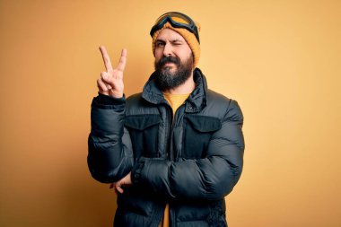 Sakallı, kar maskesi takmış, kar maskesi takmış ve kameraya gülümseyen yakışıklı, kel bir kayakçı zafer işareti yapıyor. İki numara..