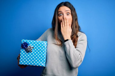 Genç, güzel esmer bir kadın, mavi arka plan üzerinde doğum günü hediyesini tutuyor. Ağzı kapalı, utancından ağzı açık, korkusunun dışa vurumu, sessizlikten korkması, gizli konsept.