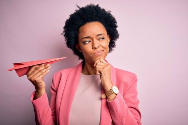 Genç Afro-Amerikalı kıvırcık saçlı, pembe kâğıt uçak tutan, ciddi yüzlü soru soran, kafası karışık bir kadın.