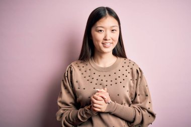 Genç, güzel Asyalı bir kadın pembe arka planda şık bir süveter giyip ellerini kavuşturuyor ve mutlu ve rahat bir şekilde gülümsüyor. Başarı ve iyimser