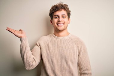 Sarışın, kıvırcık saçlı, beyaz arka planda sıradan bir süveter giyen neşeli bir şekilde gülümseyen ve avuç içi kameraya bakan genç bir adam..