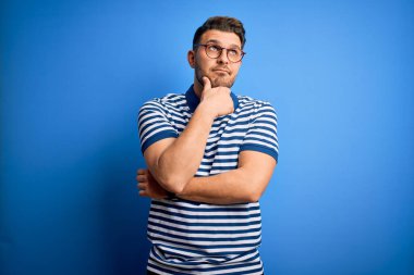 Mavi gözlü, gözlüklü ve mavi arkaplanlı sıradan çizgili tişörtlü genç bir adam. Elleri çenesinde soru sorarken, düşünceli bir ifade takınıyor. Düşünceli bir yüzle gülümsüyor. Şüphe kavramı.