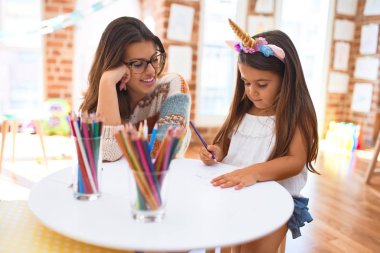 Anaokulundaki bir sürü oyuncağın etrafına kağıt ve kalem kullanarak tek boynuzlu at resmi çizen güzel öğretmen ve çocuk.