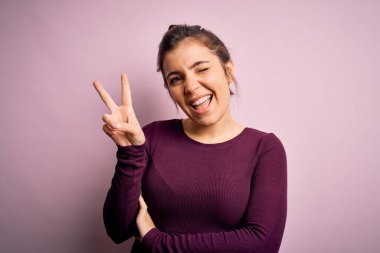 Güzel bir genç kadın pembe izole edilmiş arka planda sıradan bir çörek saç stili takıyor ve kameraya gülümsüyor zafer işareti yapıyor. İki numara..