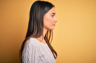 Sarı arka planda beyaz süveter giyen genç, güzel esmer kadın, yan yana bakıyor, rahat profil pozu veriyor, kendine güvenen bir gülümsemesi var..