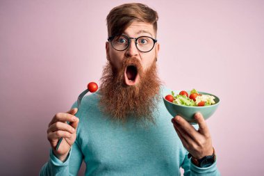 Kızıl saçlı, sakallı, pembe arka planda vejetaryen yeşil salata yiyen, korkmuş ve korku ifadesinden korkan sağlıklı bir İrlandalı.