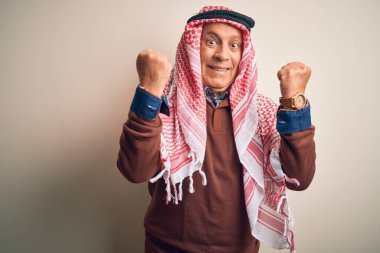 Kıdemli yakışıklı Arap adam keffiyeh giyip izole edilmiş beyaz arka planda dikilmiş kollarını kaldırarak ve açık gözlerle başarının şaşkınlığını ve şaşkınlığını kutluyor. Kazanan konsept.