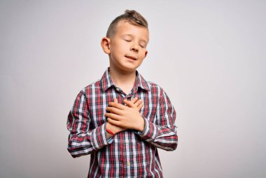 Mavi gözlü, zarif gömlekli, soyutlanmış arka planda duran, elleri göğsünde, yüzü kapalı ve minnettar bir şekilde gülümseyen küçük beyaz bir çocuk. Sağlık kavramı.