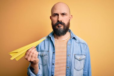 Sakallı, kel ve yakışıklı bir adam, sarı arka plan üzerinde kağıt uçak tutuyor. Zeki bir yüz ifadesi var ve ciddi düşünüyor.