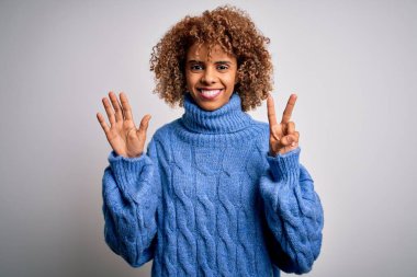 Beyaz arka plan üzerine balıkçı yaka kazak giyen güzel Afro-Amerikalı kadın yedinci parmaklarını gösterirken kendinden emin ve mutlu bir şekilde gülümsüyor..