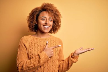 Young beautiful African American afro woman with curly hair wearing casual sweater amazed and smiling to the camera while presenting with hand and pointing with finger.