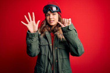 Kayak için kayak gözlüğü ve kışlık ceket giyen genç sarışın kız kırmızı arka planda ve parmaklarıyla altı numarayı gösterirken kendinden emin ve mutlu bir şekilde gülümsüyor..