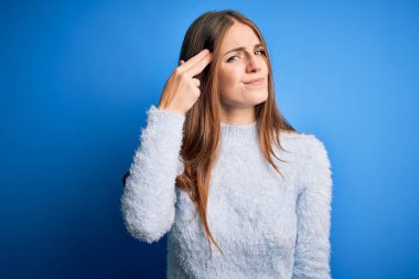 Genç, güzel, kızıl saçlı bir kadın, izole edilmiş mavi arka plan üzerine günlük süveter giyip kendini vuruyor ve kendini ve parmaklarını silah gibi havaya kaldırarak öldürüyor, intihar jesti..