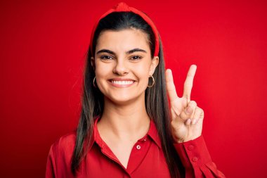 Genç, güzel, esmer bir kadın, gündelik tişört giyiyor ve kırmızı arka plan üzerinde tepe taklak, gülümsüyor, kameraya göz kırparak zafer işareti yapıyor. İki numara..