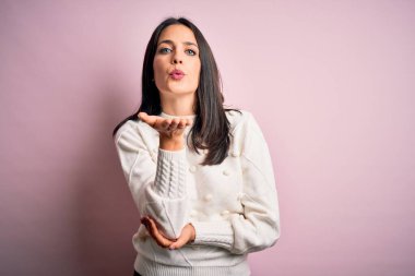 Mavi gözlü genç esmer kadın izole edilmiş pembe arka planda sıradan bir süveter giyiyor. Kameraya bakıyor ve havada el ele öpüşüp güzel ve seksi bir şekilde öpüşüyor. Aşk ifadesi.