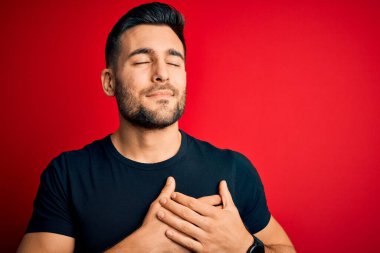 Siyah bir tişört giyen yakışıklı genç adam izole edilmiş kırmızı arka plan üzerinde duruyor elleri göğsünde, gözleri kapalı ve yüzünde minnet dolu bir jest ile gülümsüyor. Sağlık kavramı.