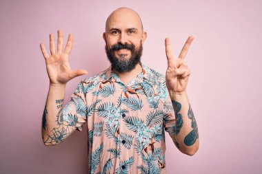 Sakallı ve dövmeli yakışıklı bir adam pembe arka planda sıradan bir çiçek gömleği giyiyor ve yedinci parmaklarını gösterirken kendinden emin ve mutlu bir şekilde gülümsüyor..