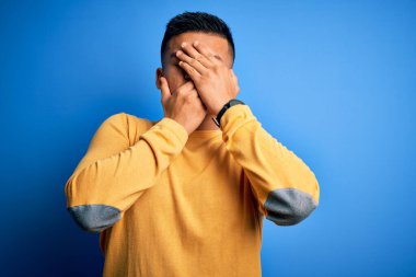 İzole edilmiş mavi arka planda sarı günlük süveter giyen genç, yakışıklı Latin adam gözleri ve ağzı kapalı, şaşırmış ve şok olmuş. Duygularını gizliyor.