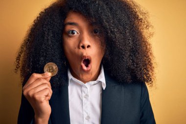 Afro saçlı, sarı arka planda bitcoin tutan Afro-Amerikalı genç bir iş kadını. Şaşırmış bir yüzle korkmuş ve korku ifadesinden heyecanlanmış.