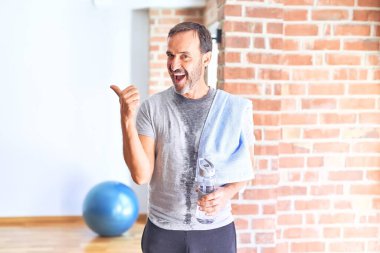 Middle age handsome sportman sweaty holding bottle of water and towel after exercise at gym pointing and showing with thumb up to the side with happy face smiling