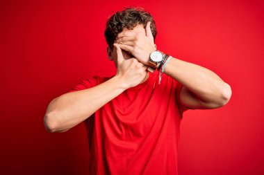 Sarışın, kıvırcık saçlı, kırmızı arka planda sıradan tişörtler giyen gözleri ve ağzı kapalı, şaşırmış ve şok olmuş genç bir adam. Duygularını gizliyor.