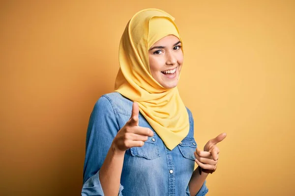 若い美しい少女は幸せと面白い顔を持つカメラに指を指して孤立した黄色の背景の上に立ってイスラム教徒のヒジャーブを着て 良いエネルギーと振動 — ストック写真