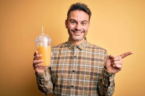 年轻英俊的男子喝着健康的橙汁 用稻草盖住黄色的背景 非常高兴地将手和手指指向旁边 — 图库照片